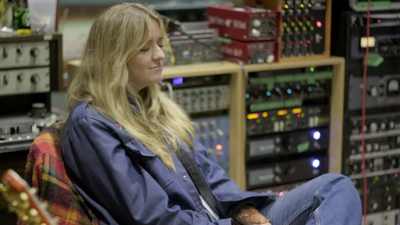 Blonde-haired Female Artist Sitting In Recording Studio And Vibing To The Beat, With Audio Equipment In Background. medium shot
