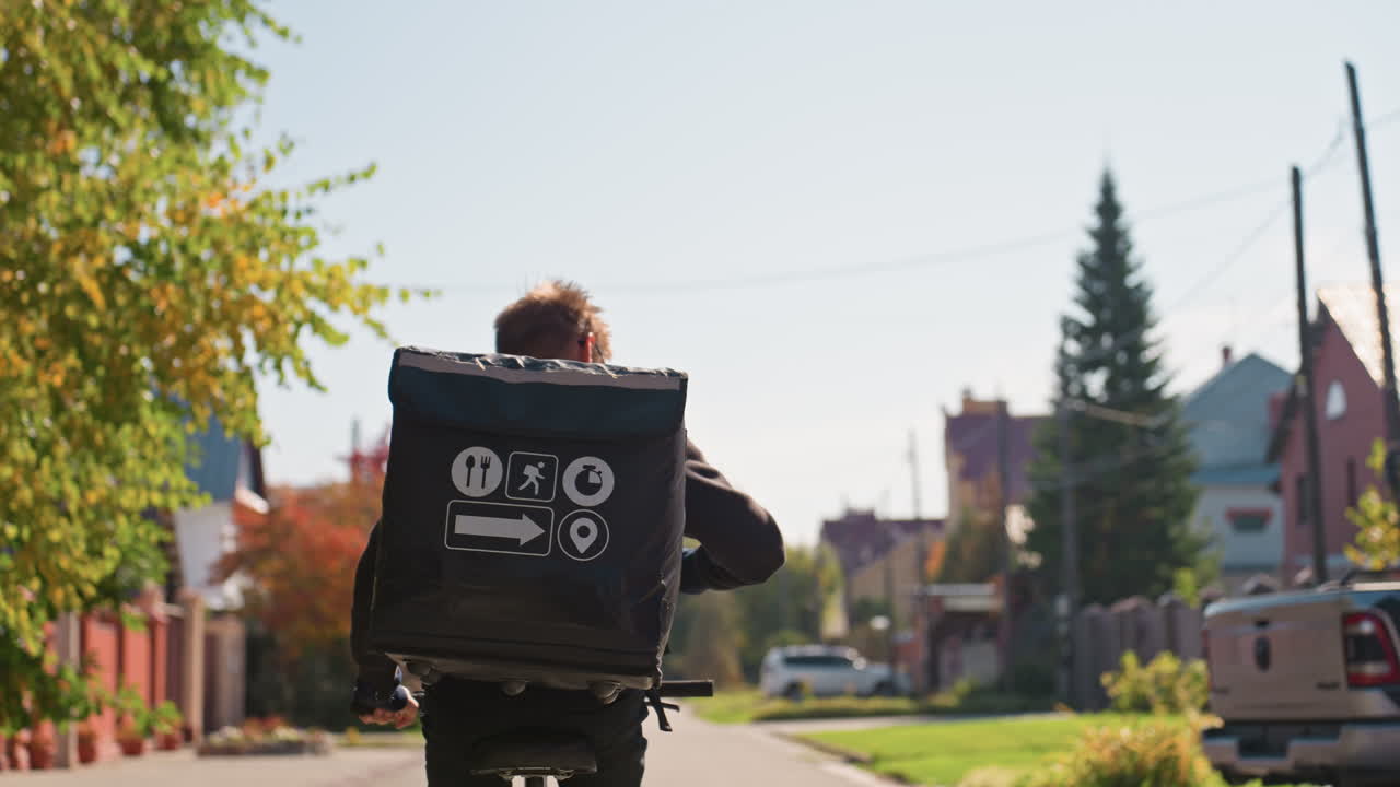 Caucasian courier cycling suburban street with insulated backpack and branded icons. Morning sunlight warms treelined residential road with colorful autumn foliage. Rider navigates quiet lanes