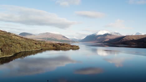 Aerial-drone-footage-flying-backwards-above-Loch-Etive-facing-Glen-Etive-in-the-Highlands-of-Scotland-in-winter-with-snow-capped-mountains,-trees-and-native-forest-with-blue,-reflective,-still-water