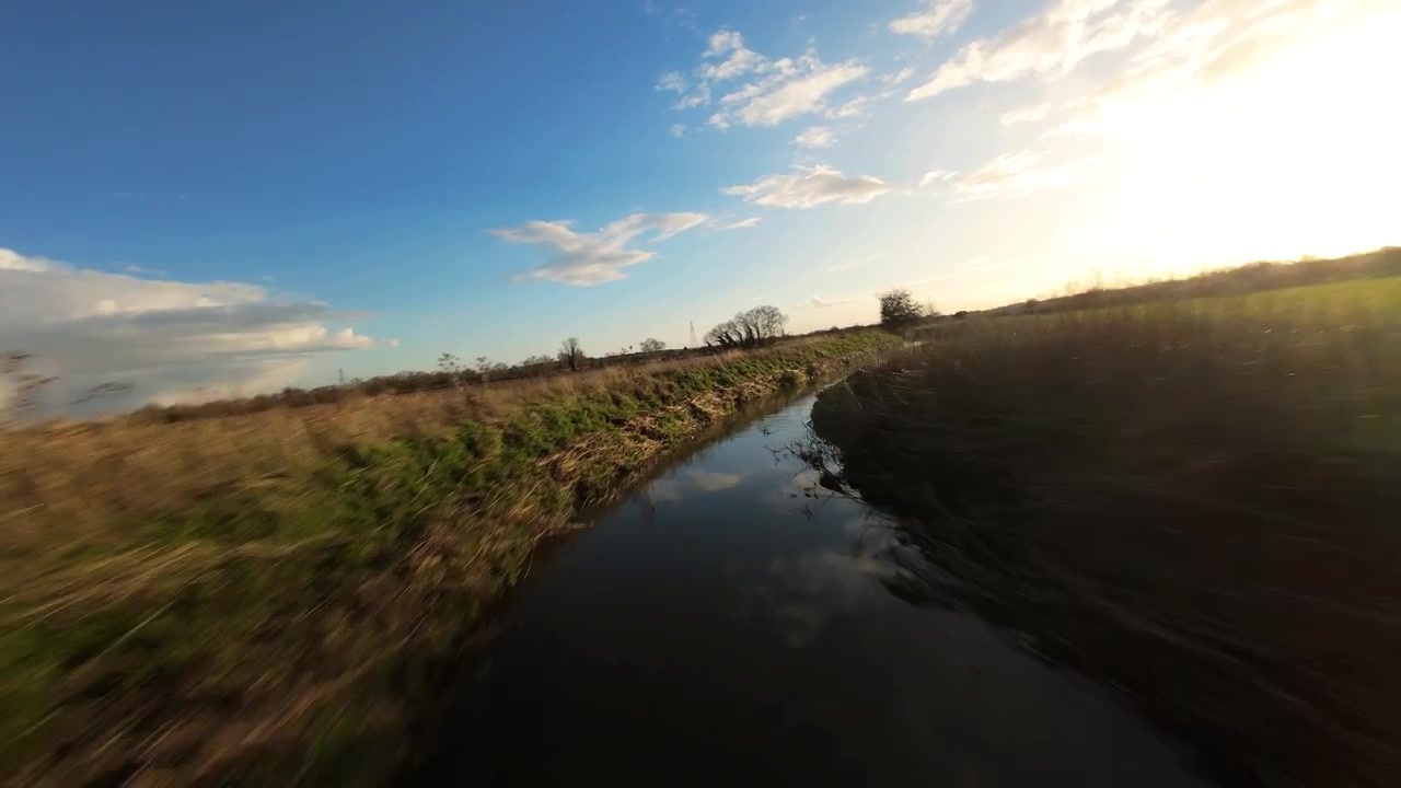 Ripresa veloce con drone FPV a bassa quota che vola lungo un argine di drenaggio stretto, attraversando vivaci pascoli agricoli verdi sotto un cielo arancione drammatico.