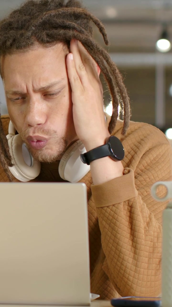Vertical video: Reacting to laptop, dreadlocked man in sweater rubbing jaw at desk, with headphones