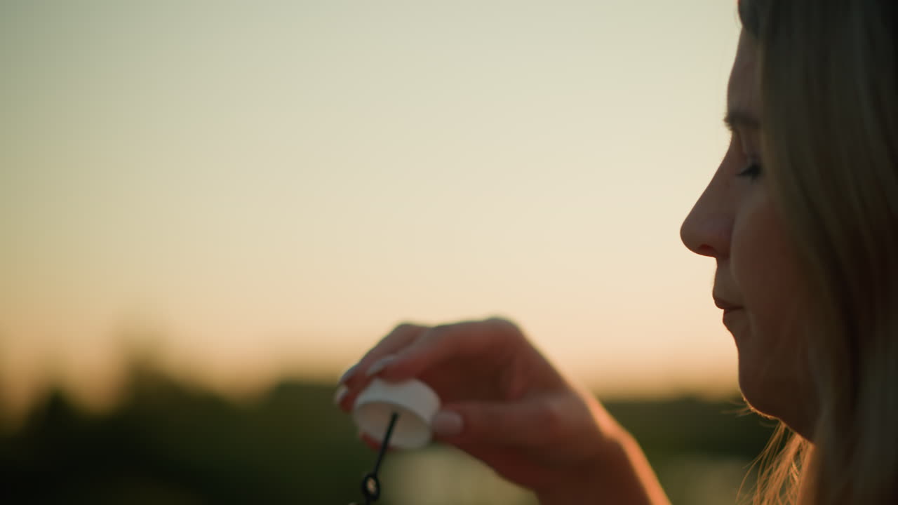 close-up de mulher branca soprando bolhas ao ar livre durante o pôr-do-sol com óculos de sol na cabeça, a luz solar ilumina suavemente seu rosto enquanto ela segura a varinha de bolhas