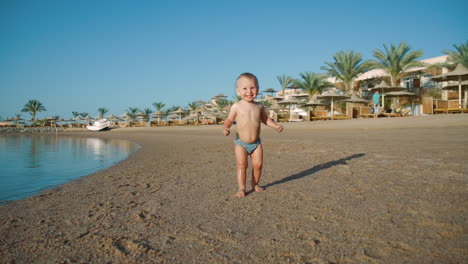 Joyful-baby-boy-walking-along-sand-coastline-to-water-at-summer-day.
