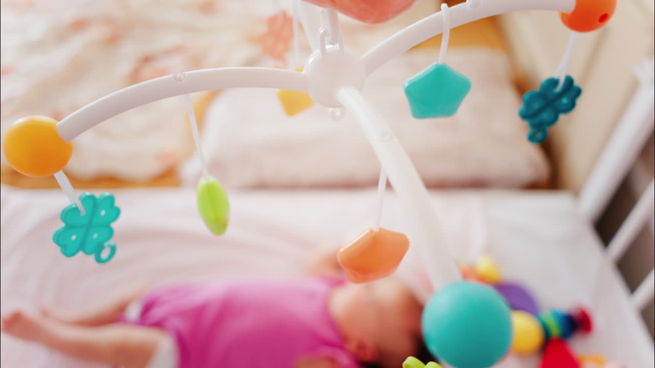 Baby lying on a bed in a pink outfit, looking up at a colourful hanging mobile toy