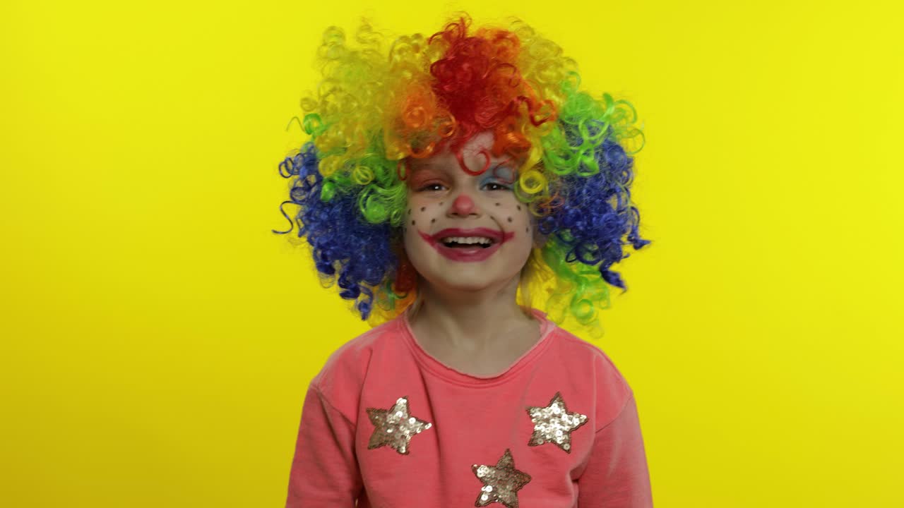 niña payasa en peluca arco iris haciendo caras tontas. divirtiéndose, sonriendo. pulgar hacia arriba. halloween