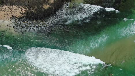 Burleigh-Heads,-Surf-Rolling-into-Shore,-Rocks,-Surfers,-Above-AERIAL