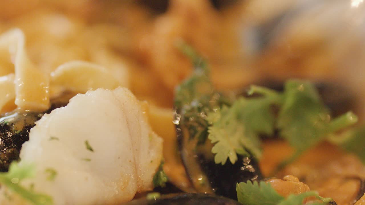 Macro view of assorted seafood including fish, squid, scallop, and mussels in a hot chowder, garnished with cilantro. Warm lighting, shallow depth of field, and subtle camera movement highlight textures and freshness
