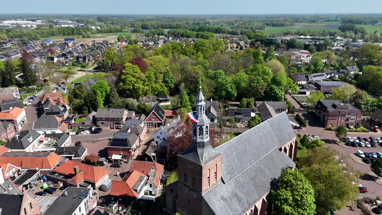 Historic church of Groenlo with waving durch flag on sunny day. People on streets celebrating Kings Day in Netherlands. Spring day with green and red colored trees. Aerial orbit shot.