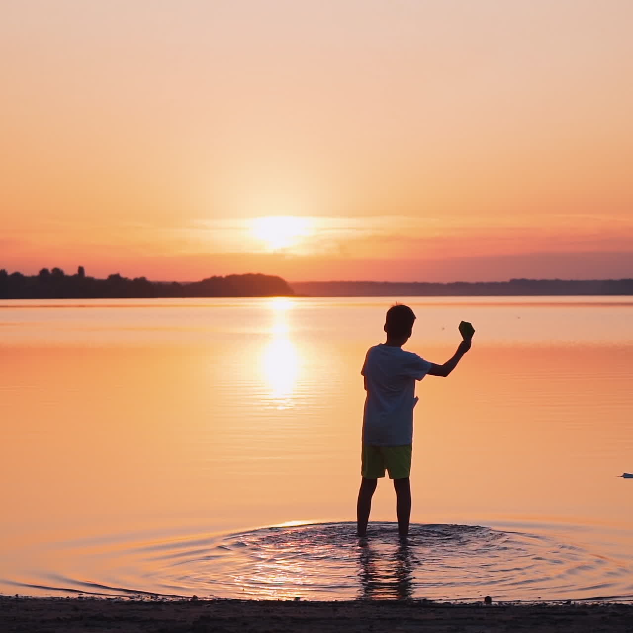 Backside view of a boy in the evening. Child standing in water and launching paper plane into the river at sunset. Slow motion.
