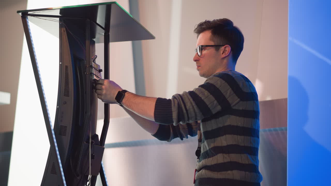 man adjusts back of digital display screen in video production studio, focused on setup and connection, wearing striped sweater and ID badge, with blue lighting and modern equipment backdrop