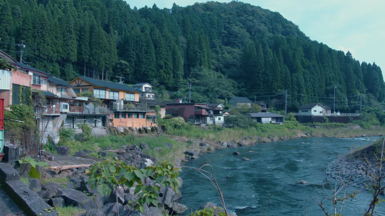 escena pacífica de un remoto pueblo japonés a orillas del río rodeado de bosque - vista estática en kirishima, japón