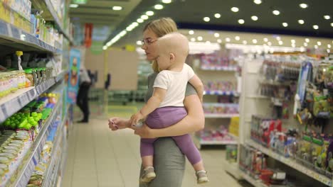Beautiful-woman-in-glasses-holding-her-child-in-her-arms-while-choosing-baby-food-on-the-shelves-in-the-supermarket.-fruit-puree
