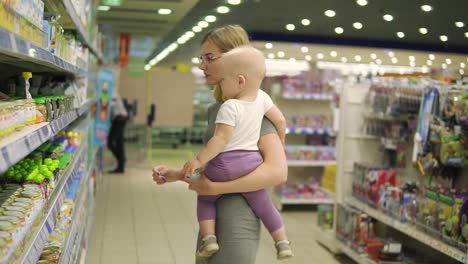 hermosa mujer con gafas sosteniendo a su hijo en sus brazos mientras elige comida para bebés en los estantes del supermercado. puré de frutas