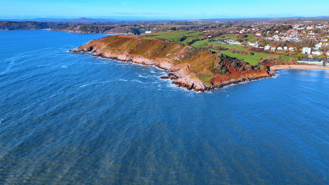 Vista drone della costa di Langland Bay e della zona residenziale. Prospettiva aerea che mostra la transizione dalle scogliere rocciose e i campi verdi a un villaggio costiero in Galles.