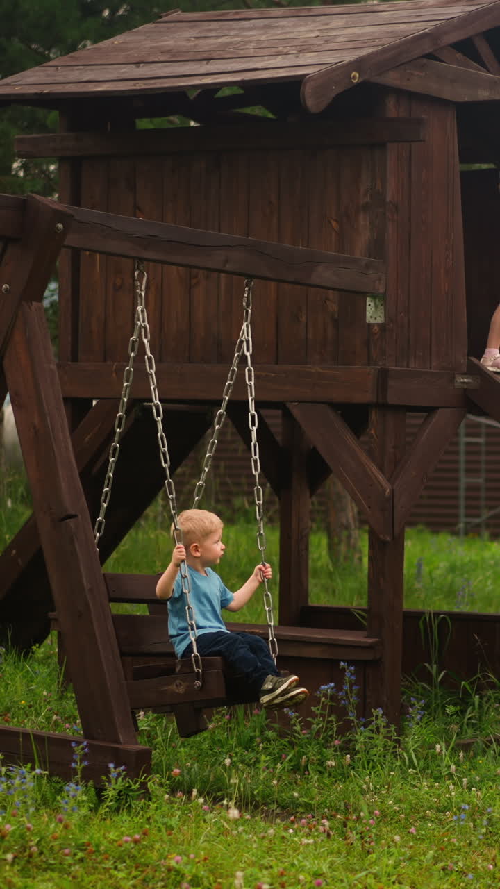 Little girl points ahead by finger standing on wooden children house slide and younger brother swings on playground at eco resort in forest slow motion