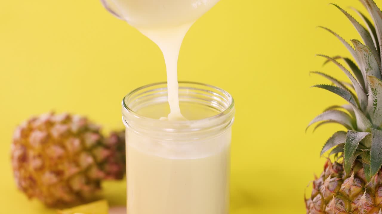 Pineapple smoothie poured into glass jar, bright yellow background, vibrant lighting, static camera angle