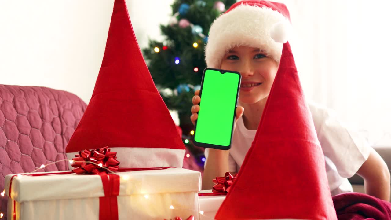 산타클로스 모자를 입은 행복한 웃는 소년은 초록색 배경으로 휴대전화를 들고 아한 크리스마스 인테리어에서 소파에 앉아 있습니다. 녹색 화면 셀