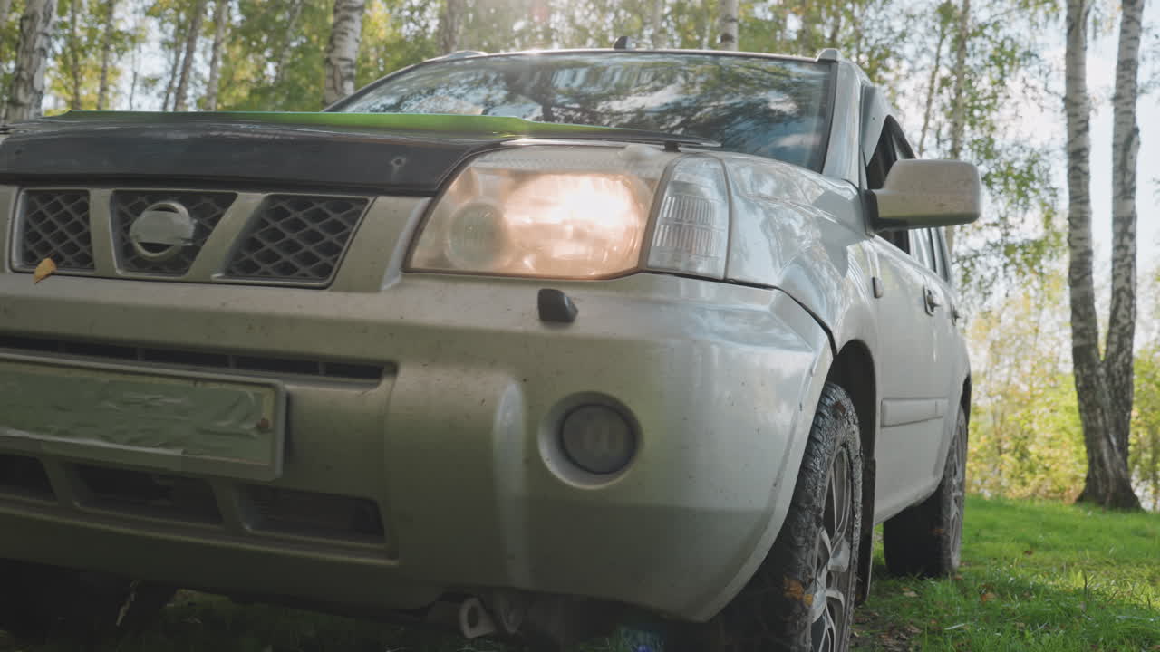 Campeggiatore esce dal SUV in una radura sul lago, luce solare retroillumina figura in giacca gialla, persona che cerca l'attrezzatura, apre la porta, vetro riflettente e tronchi di betulla incorniciano la scena, atmosfera calda e accogliente all'aperto