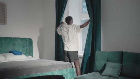 man in white t-shirt standing in minimalist bedroom, pulling dark green curtains apart to let sunlight fill room, with green headboard and neatly made bed visible in serene atmosphere