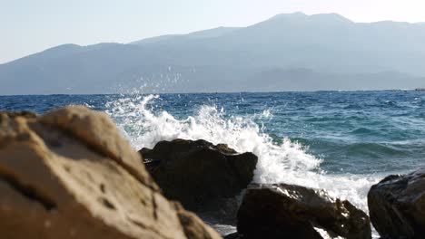 Primer-Plano-De-Grandes-Rocas-En-La-Playa-Albanesa-Destrozadas-Por-Las-Olas-Azules-Del-Mar-Jónico,-Con-Hermosas-Montañas-Al-Fondo