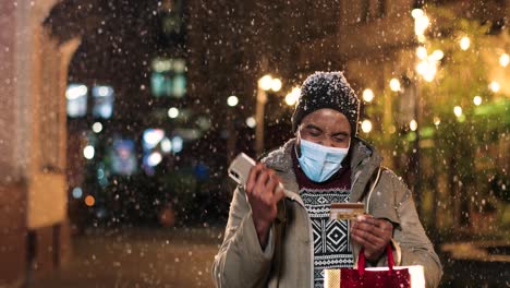 Retrato-De-Un-Joven-Afroamericano-Con-Máscara-Facial-Escribiendo-En-Un-Teléfono-Inteligente-Y-Comprando-En-Línea-En-La-Calle-Mientras-Nieva-En-Navidad