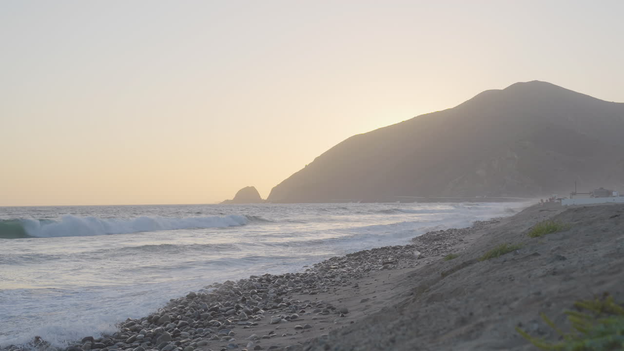 toma estacionaria en cámara lenta de olas corriendo a lo largo de las orillas de la playa de mondo mientras el sol se pone detrás de la montaña ubicada en el sur de california