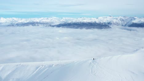 Solo-person-waits-on-top-of-snow-covered-mountain-ridge-above-sea-of-clouds,-aerial-orbit