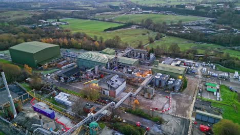 Slow-forward-moving-aerial-drone-footage-of-a-small-industrial-plant-at-dusk,-showing-pipework-structures,-buildings,-cooling-towers,-steam,-and-work-vehicles