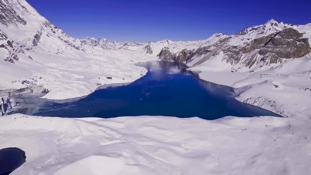 Aerial drone footage flying lower and closer to blue Tilicho lake in Nepal during a sunny day. The lake is surrounded by white covered snowy land and snow covered mountains. Blue sky in the background