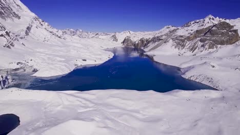 aerial drone footage flying lower and closer to blue tilicho lake in nepal during a sunny day. the lake is surrounded by white covered snowy land and snow covered mountains. blue sky in the background