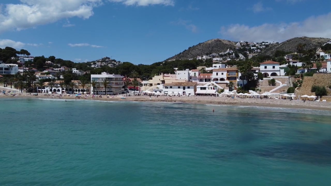 Cámara de dron volando rápido a baja altitud sobre aguas cristalinas del océano hacia la playa de El Portet en Moraira, España. Toma de la soleada costa mediterránea
