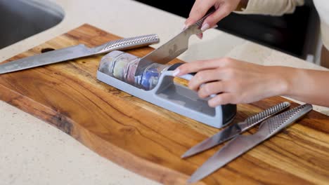 A person sharpens a knife using a manual sharpener on a wooden cutting board in a well-lit kitchen