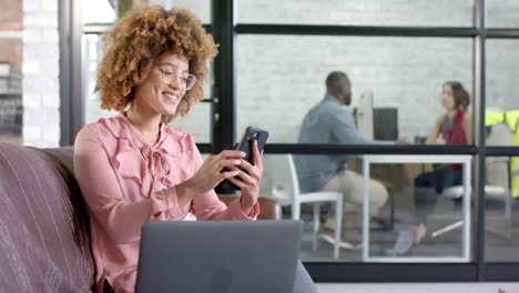 feliz mujer de negocios casual biracial usando smartphone en el sofá en la oficina, espacio de copia, cámara lenta