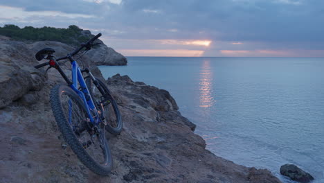 Lapso-De-Tiempo-De-La-Bicicleta-Abandonada-Apoyada-Contra-Los-Acantilados-Mientras-El-Sol-De-La-Mañana-Se-Eleva-Sobre-El-Horizonte,-Arrojando-Sus-Rayos-Al-Mar