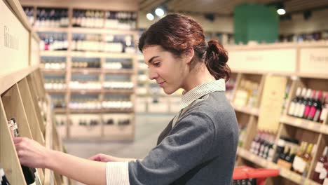 Young-beautiful-brunette-caucasian-girl-compares-two-different-bottles-of-wine.-Choosing-white-wine-at-grocery-store