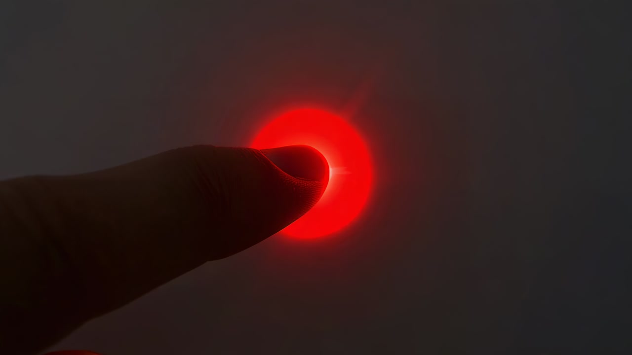 Approaching fingertip pressing and covering small red LED on dim panel, creating halo and flare