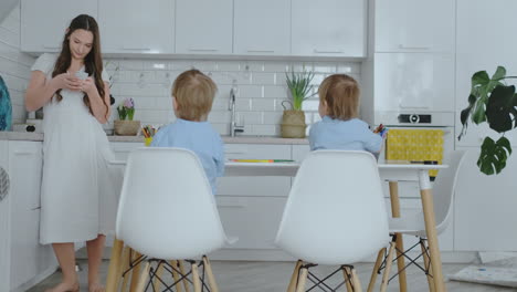 Dos-Hijos-Con-Camisas-Azules-Sentados-En-La-Mesa-Y-Dibujando-Lápices-Sobre-Papel-Sentados-En-Una-Mesa-Blanca,-Y-Mi-Madre-Escribe-Un-Mensaje-De-Texto-En-El-Teléfono-Inteligente