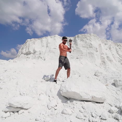 sportsman with huge hammer outdoors. sporty man demolishing big white rock by metal tool on the top of mountain in summer day. slow motion.