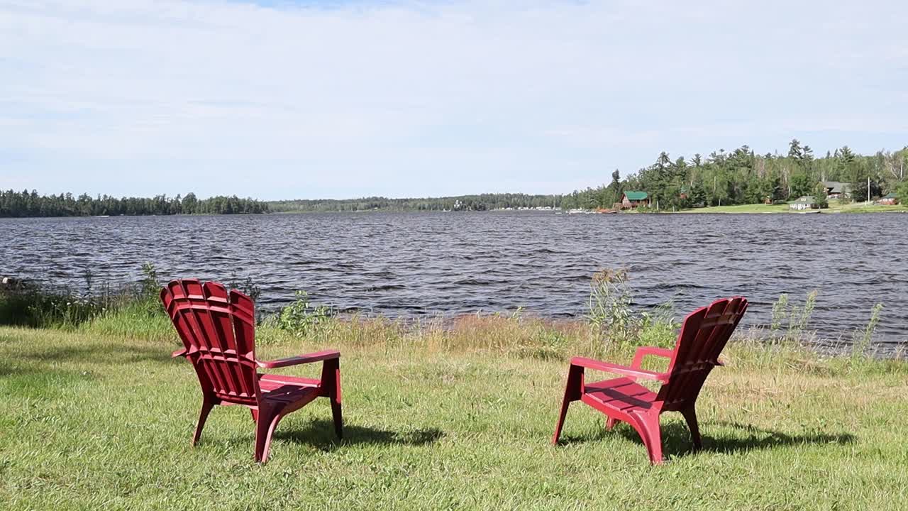 Two red Adirondack chairs sit on the shoreline at Caliper Lake in Northwest Ontario, Canada. Waves move across the lake with green grass and boreal forest trees under cloudy summer skies