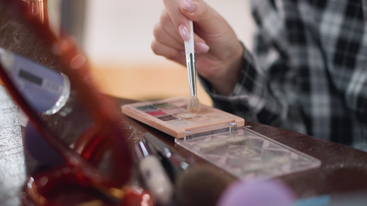 Close-up shot of lady hand holding makeup brush as she taps two different contour powders in makeup palette with blurred view of mirror in background, makeup and beauty routine concept