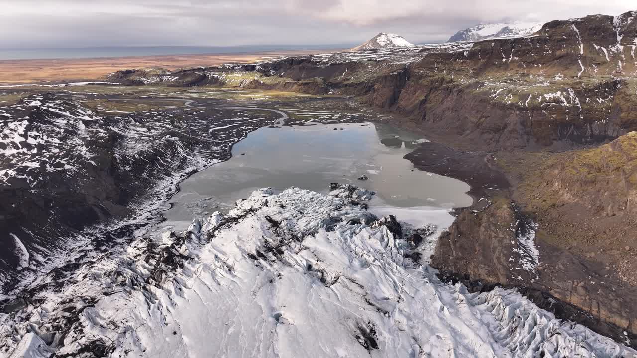 Aerial view of the glacial tongue of Sólheimajökull, Iceland, meeting a milky glacial lagoon. Rugged landscape showing ice formations and meltwater.