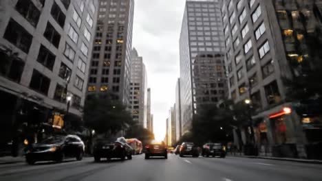 buses and cars navigate a bustling city street during evening rush hour. the sun sets between skyscrapers, creating a vibrant skyline. people move along the sidewalks.