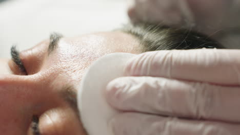close up of spa guest forehead being gently rubbed with soft cotton pad by gloved beauty technician during facial treatment in bright clinical setting with ambient lighting and serene atmosphere