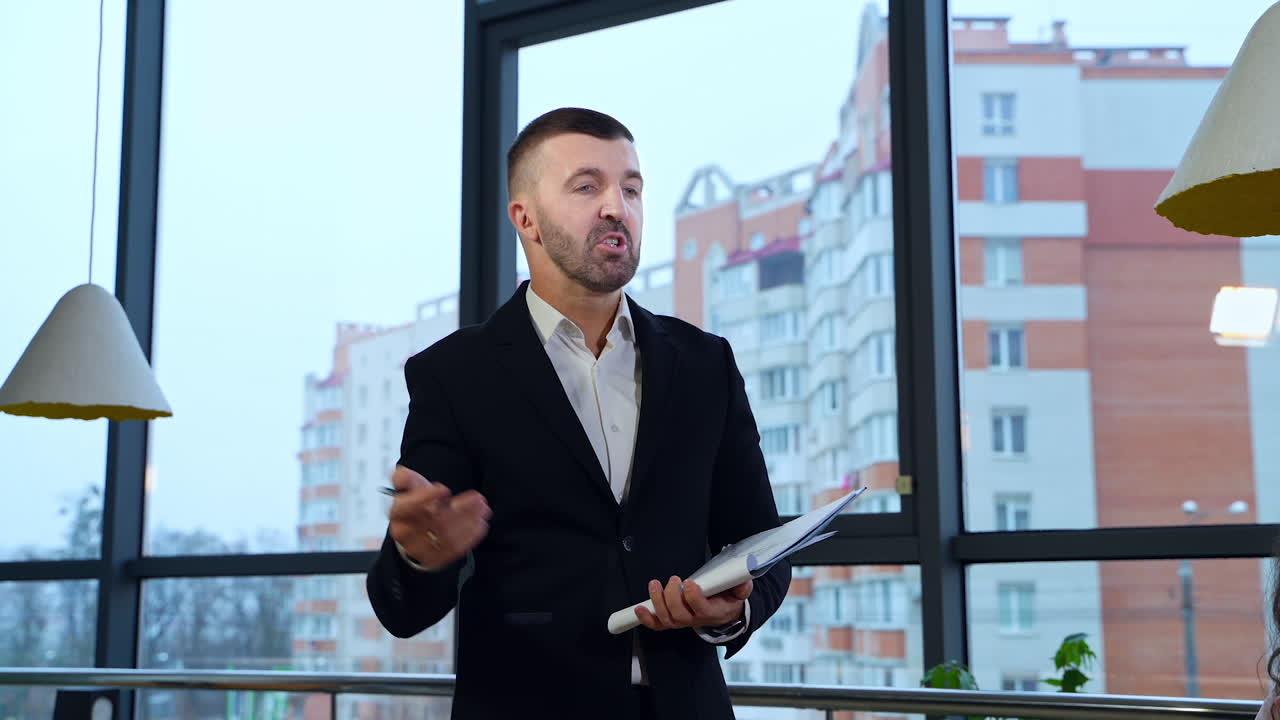 Middle-aged boss in suit standing in front of employees and holding papers. Man replying questions of his staff against city backdrop.