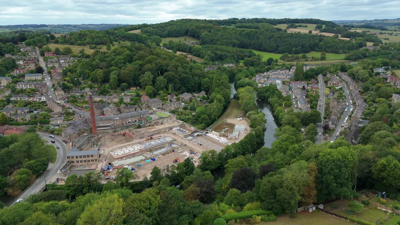Drone flight along river and through small rural town surrounded by lush green hills, Derbyshire Dales, United Kingdom