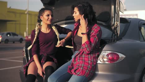 Two-Young-Attractive-Friends-Sitting-In-Car-Trunk-In-The-Parking-By-The-Shopping-Mall-And-Talking,-Smiling-Happily