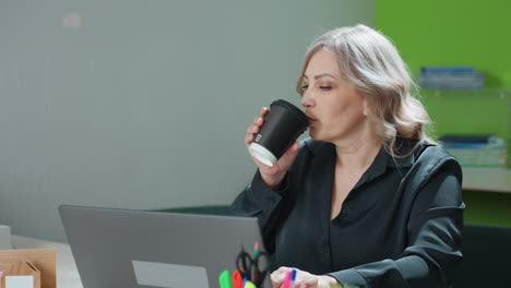 remote worker takes short pause to sip hot latte from paper cup before continuing typing, showing dedication and calm multitasking approach in home office with natural lighting and casual attire