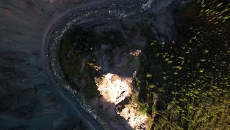 spectacular view directly above yukon burwash creek white river rapid waters flowing around curve with dense green tree forest and bright sunlight on brown dirt ground, canada, rising aerial