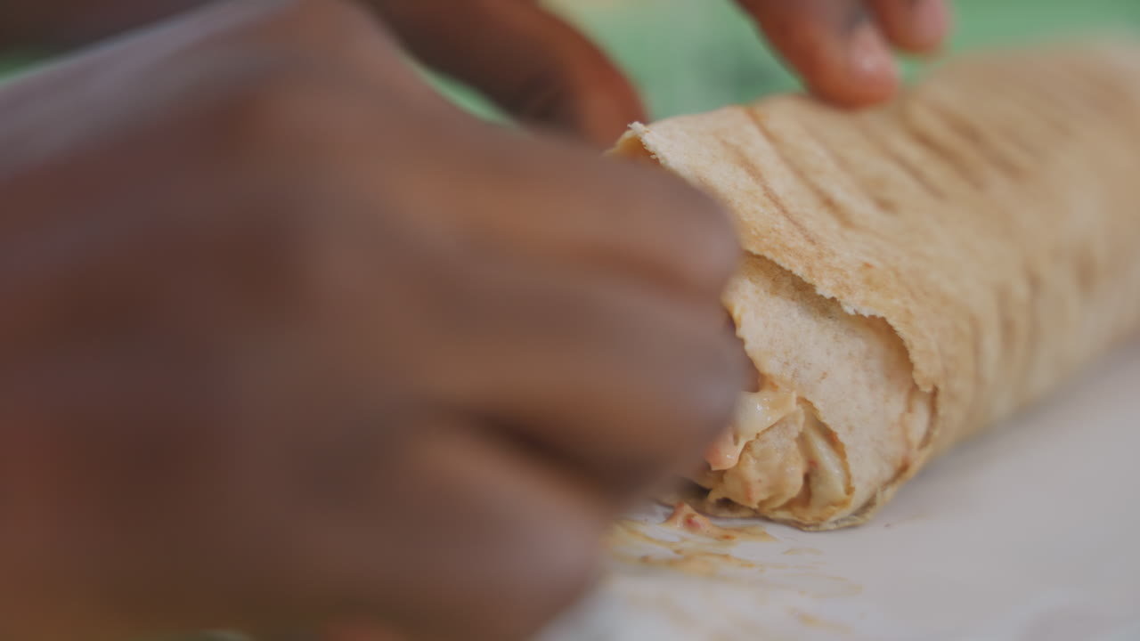 Nahaufnahme der Hände eines schwarzen Mannes, der Shawarma an einem Date-Abend hält, Finger schälen Fladenbrot, zarte Hähnchenstücke, würzige Soße, grüner Tisch, Papierserviette, entspannte Café-Atmosphäre, warmes Stadtlicht, spielerische Geste.