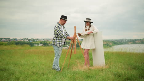 a smiling woman stands in a grassy field with her husband. the man, wearing a black hat and plaid shirt, sets up an easel while the woman, dressed in a white dress and black hat, holds a paint palette